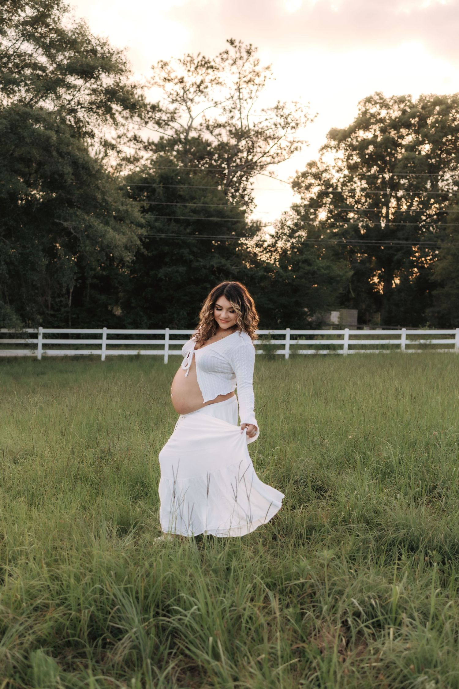 A figure in a flowing white dress twirls gracefully in a sunlit field with a white fence and trees in the background.