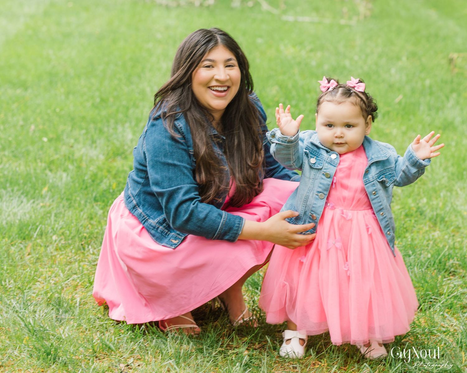 Two people in matching denim jackets and pink tulle skirts share a joyful moment on green grass outdoors.
