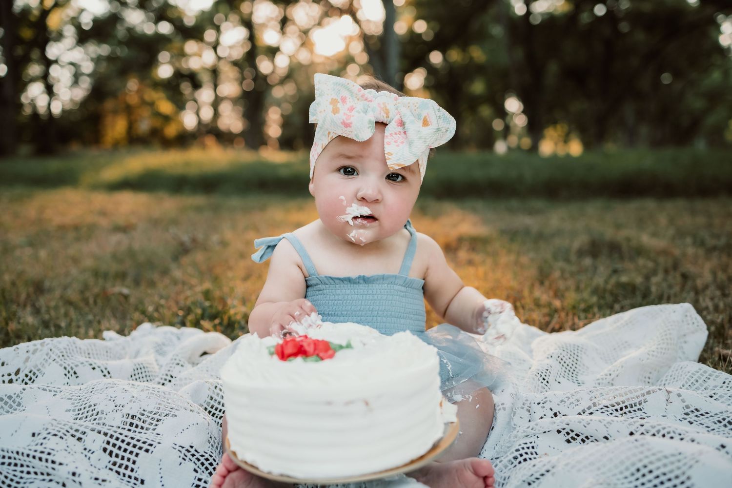 Baby in blue dress sits in grass with smashed white cake during first birthday cake smash photoshoot at sunset.