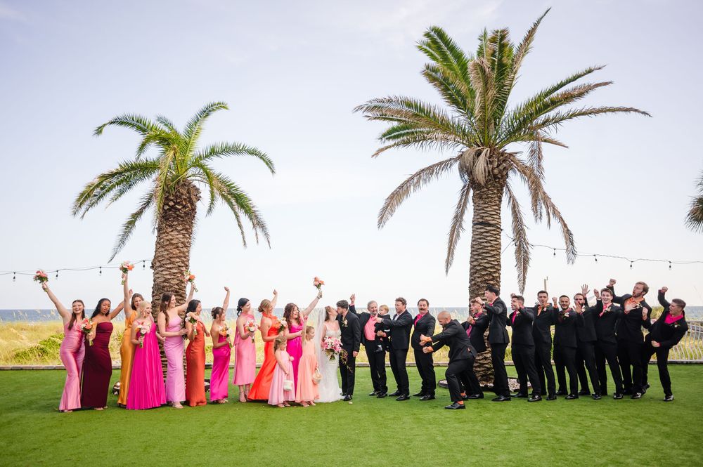 Large wedding party group strikes fun poses outdoors with palm trees and ocean in the background.