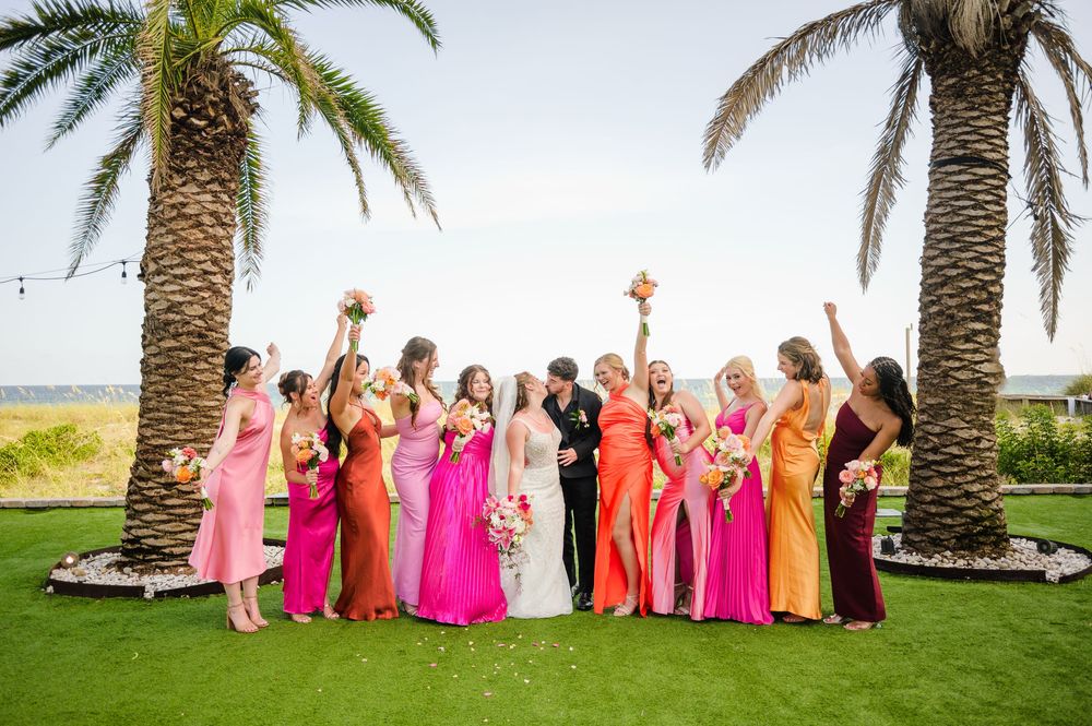 Wedding party in pink and coral dresses poses playfully together under palm trees by the ocean.