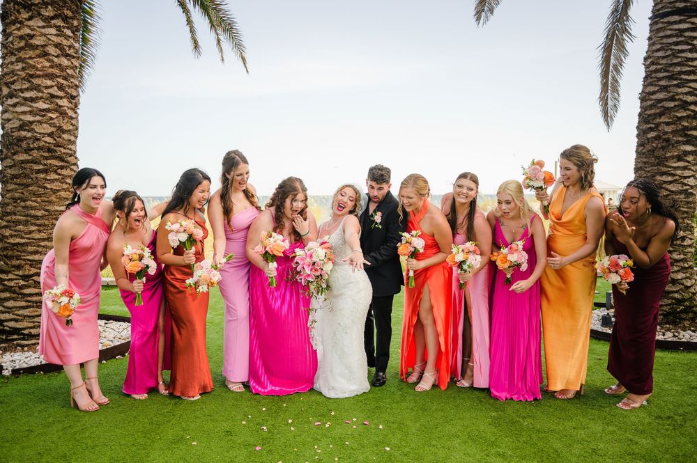 Wedding party gathered on green lawn with palm trees in colorful attire.