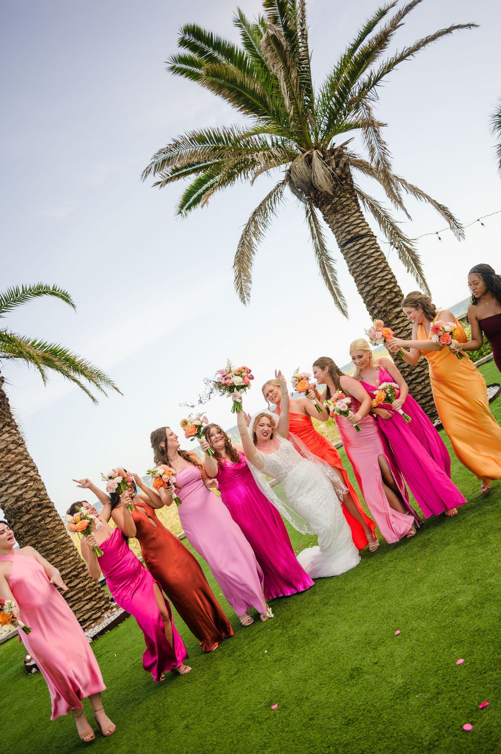 Wedding party poses under palm trees on green lawn wearing vibrant pink and orange gowns.