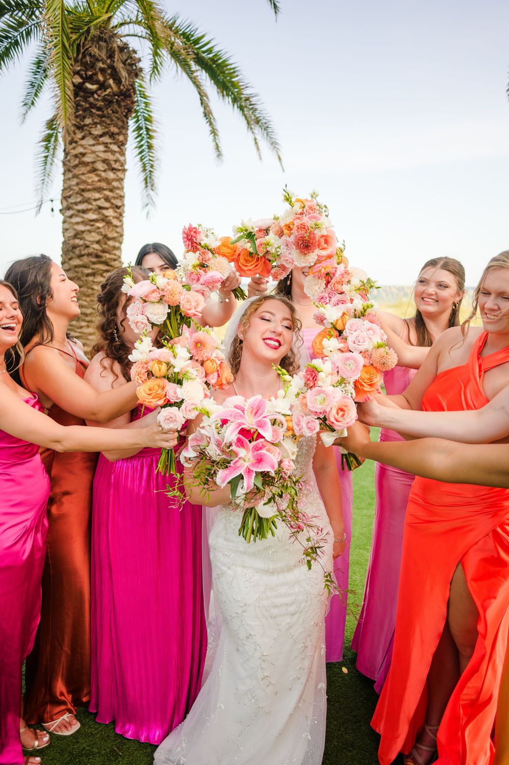 Joyful bridal party celebrates under a palm tree with colorful bouquets and bright flowing dresses.