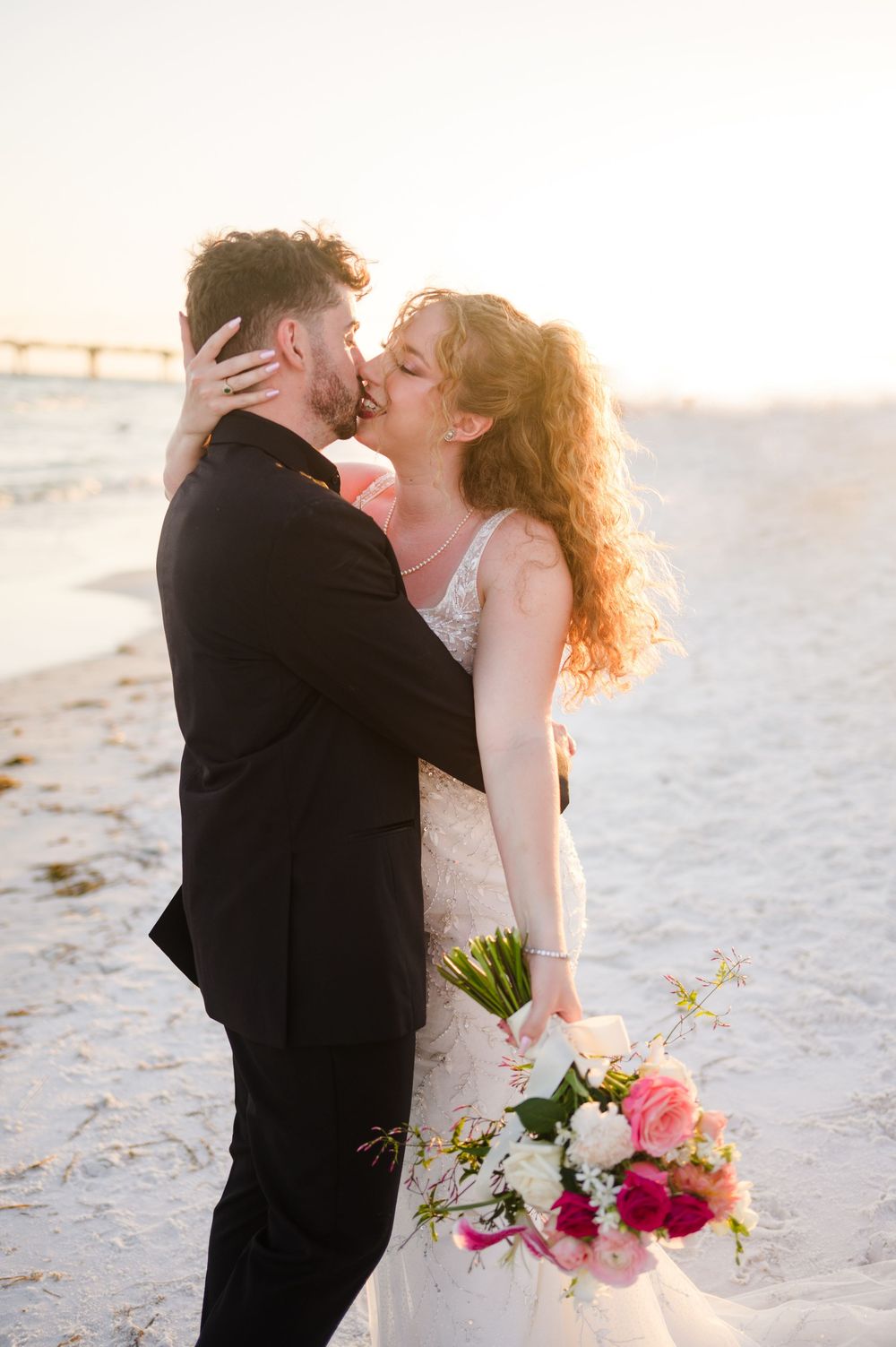 Romantic sunset beach wedding portrait with couple embracing as waves crash in background.