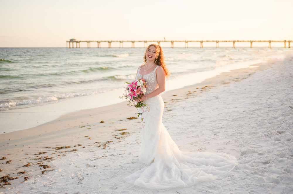 Bride in white lace mermaid gown poses with bouquet on beach at sunset with pier in background.
