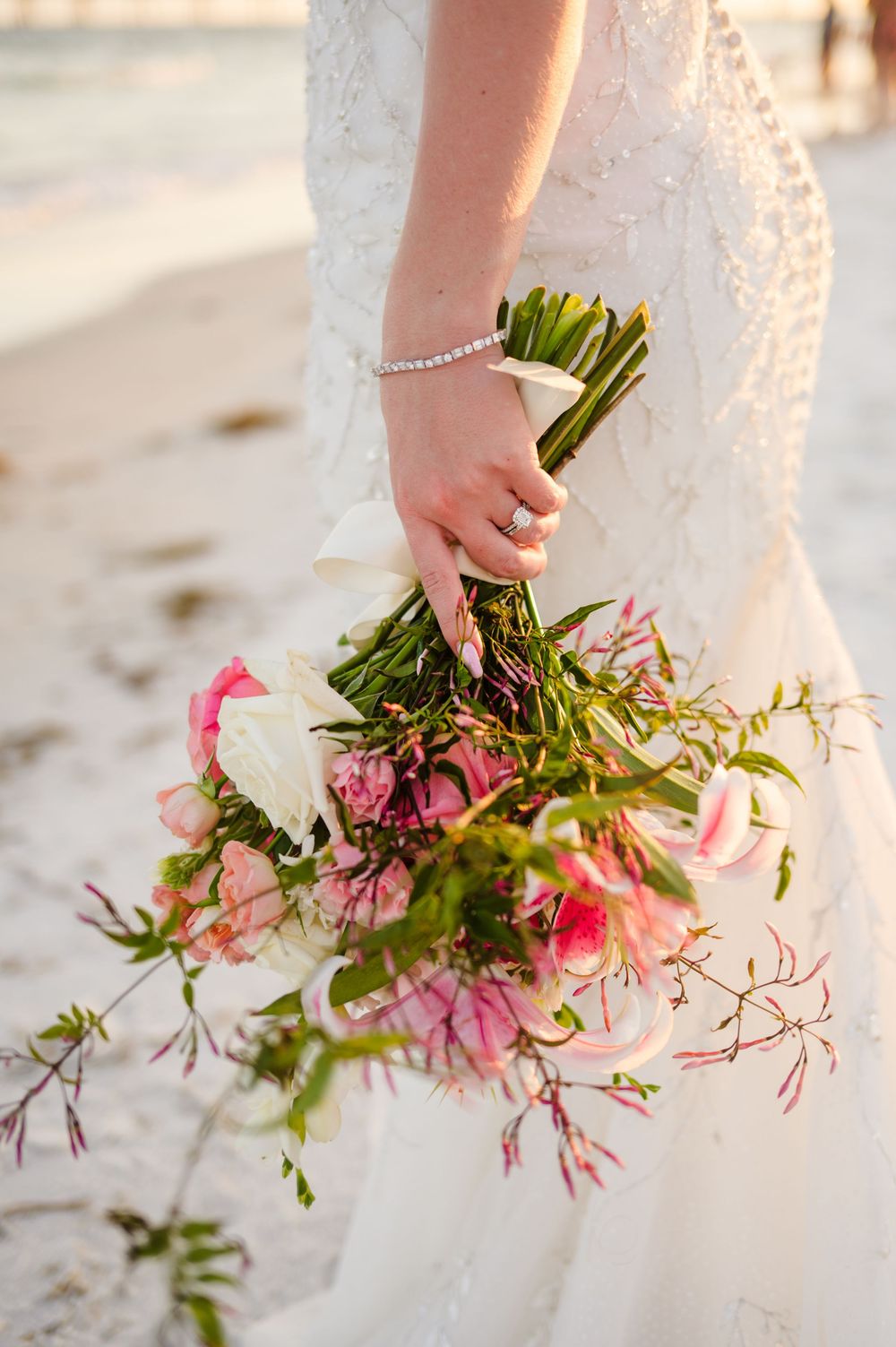 Close-up of bridal bouquet with pink and white flowers held against a lace wedding dress on beach.