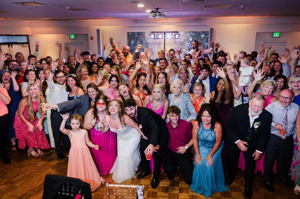 Large group of wedding guests celebrating on dance floor with colorful lighting.