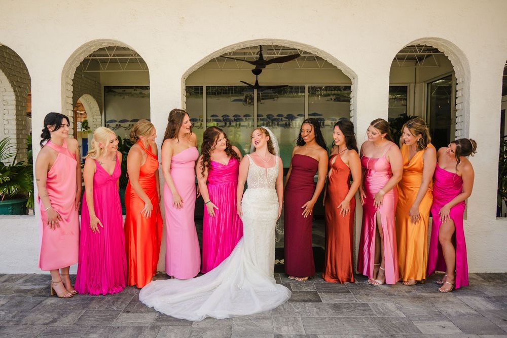 A group wearing vibrant pink and orange bridesmaid dresses stands together in an outdoor archway with a wedding gown.