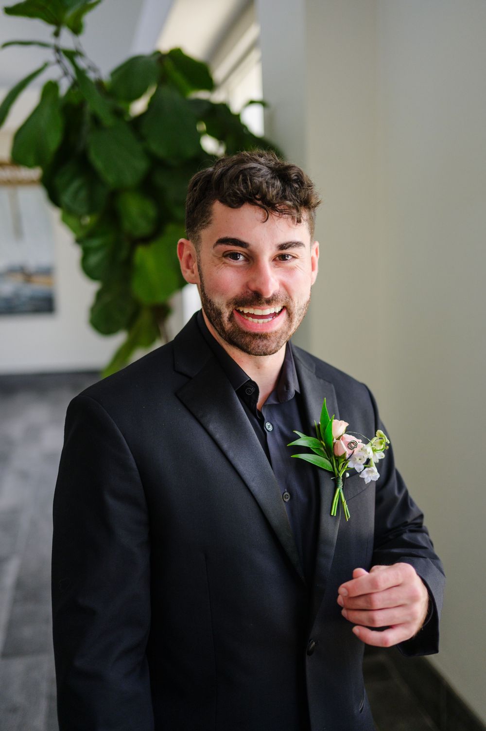 Person in formal black suit wearing a floral boutonniere smiles while standing next to indoor greenery.
