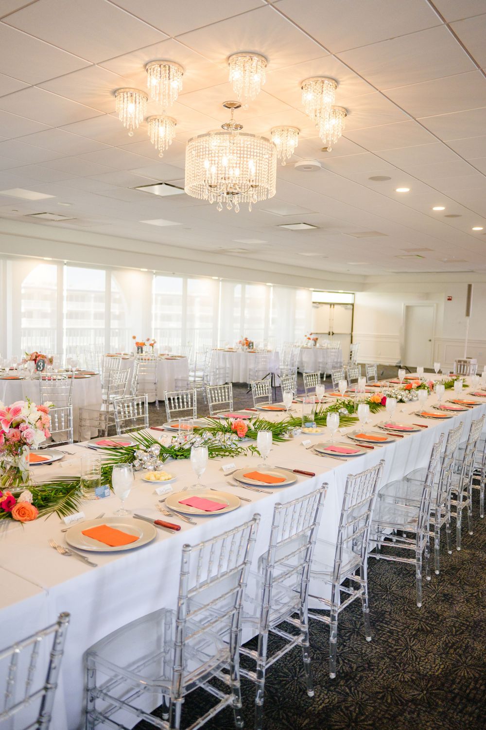 Elegant wedding reception table setting with crystal chandelier, white linens, and coral accents in a bright banquet room.
