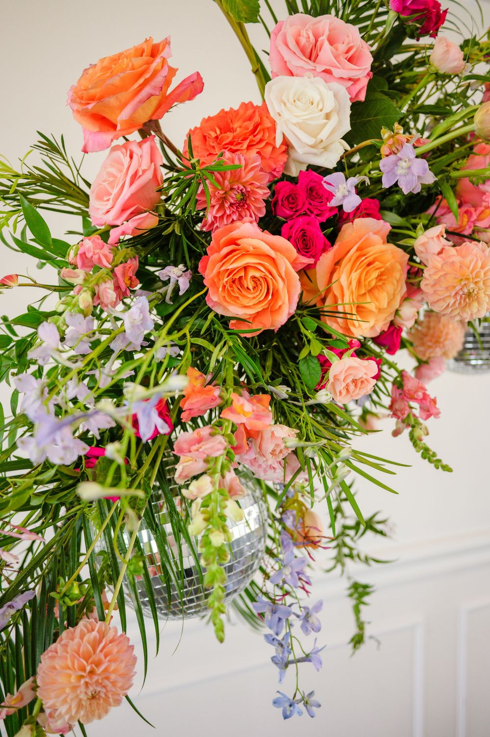 Vibrant floral arrangement with coral roses, pink carnations and purple delphinium in a glass vase with cascading greenery.
