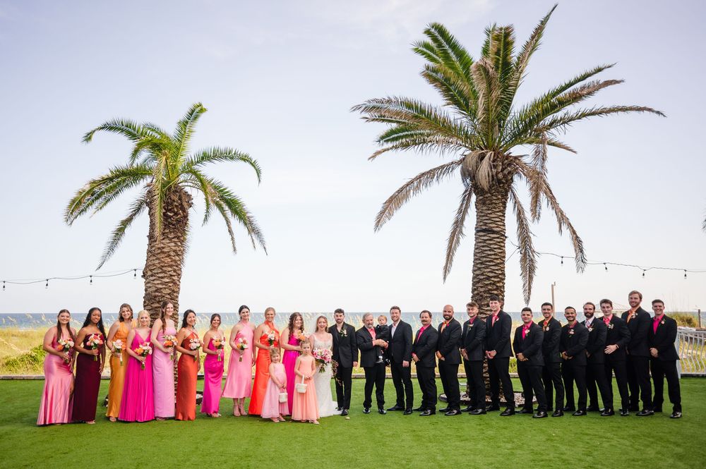 A bridal party in pink dresses and dark suits poses outdoors between two palm trees at a tropical beach venue.