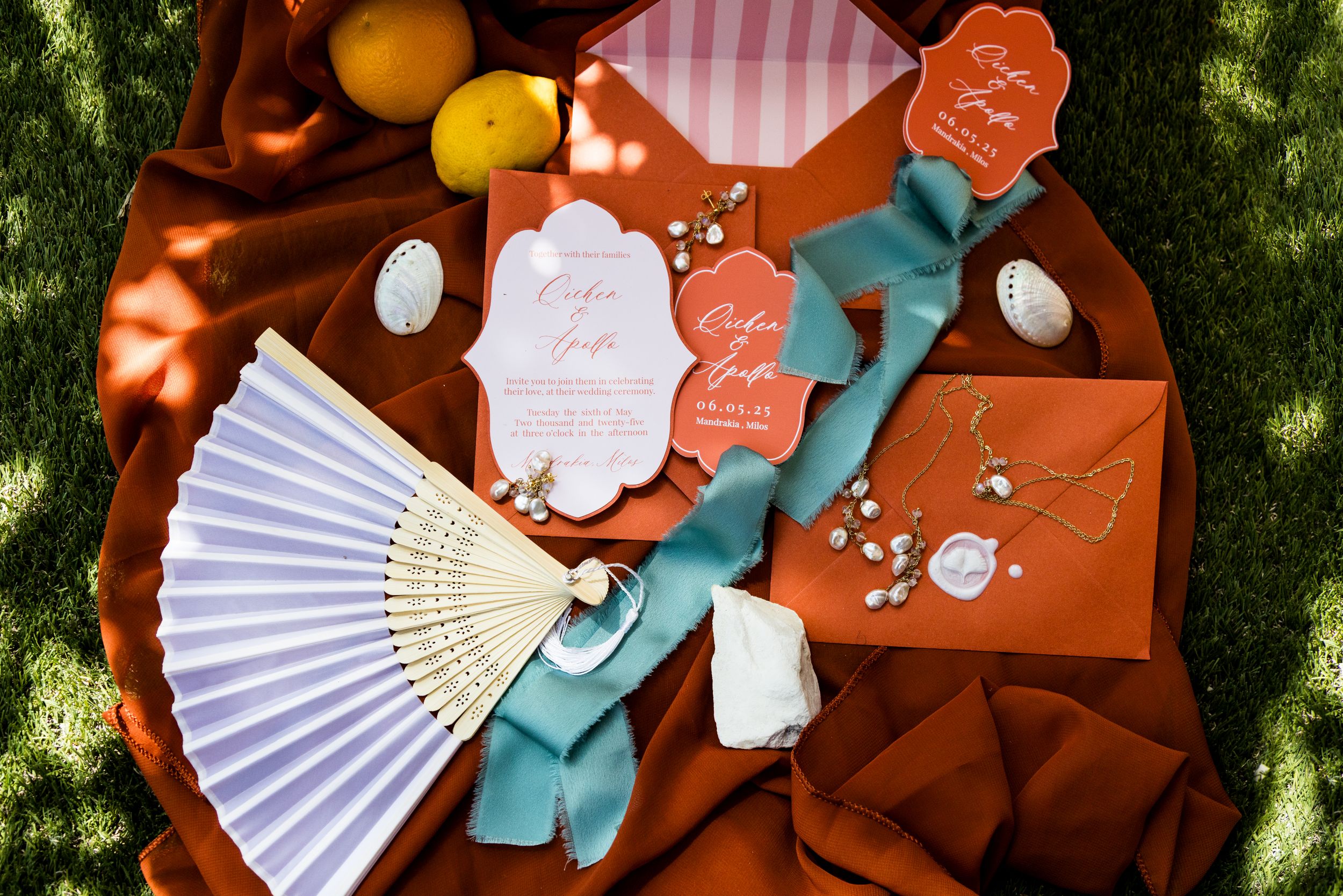 Flat lay of wedding invitations with orange envelopes, white fan, teal ribbon, and citrus fruits on terracotta fabric.