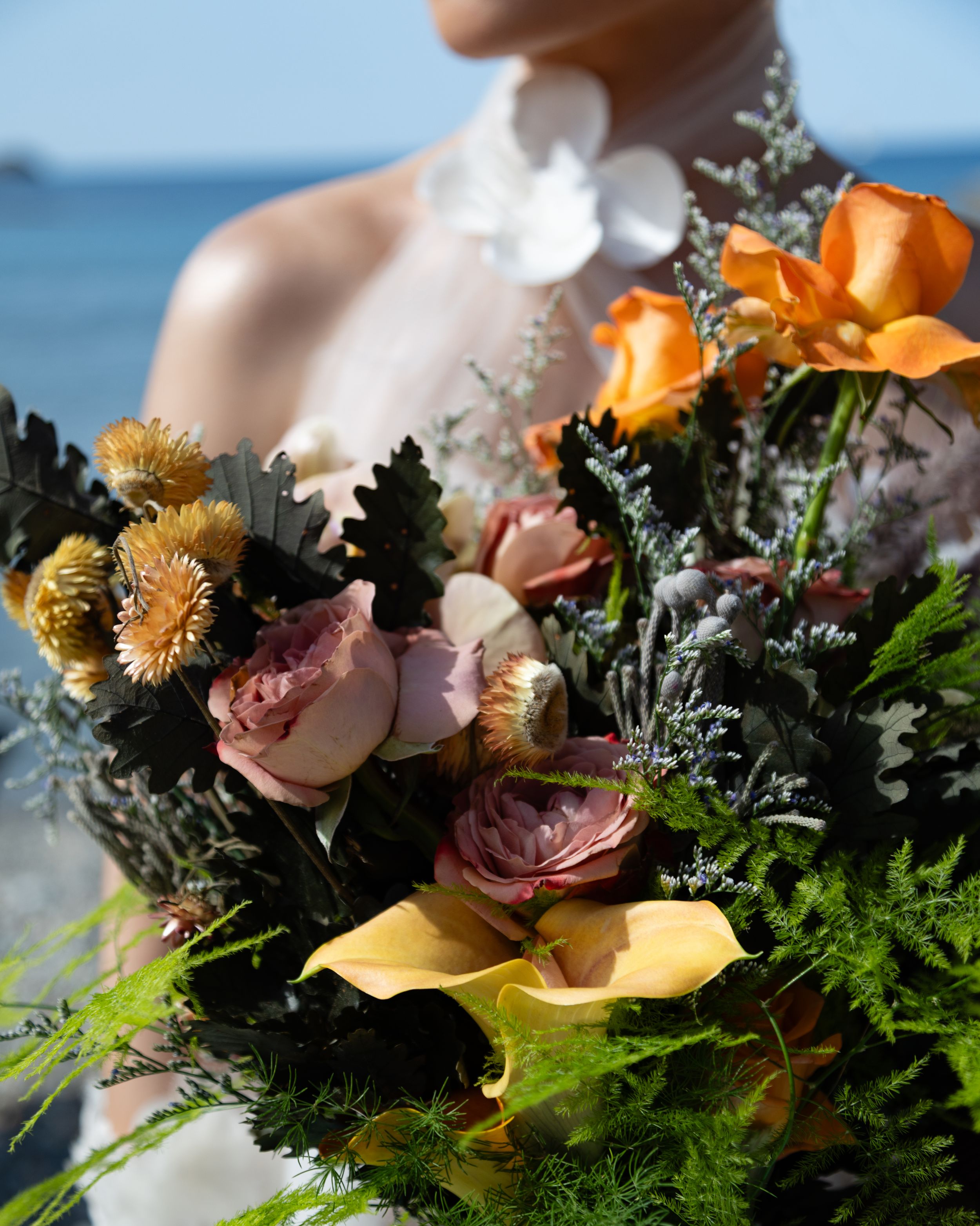 Detailed close-up of a rustic wedding bouquet with orange calla lilies, pink roses, and dried flowers against a beach backdrop.