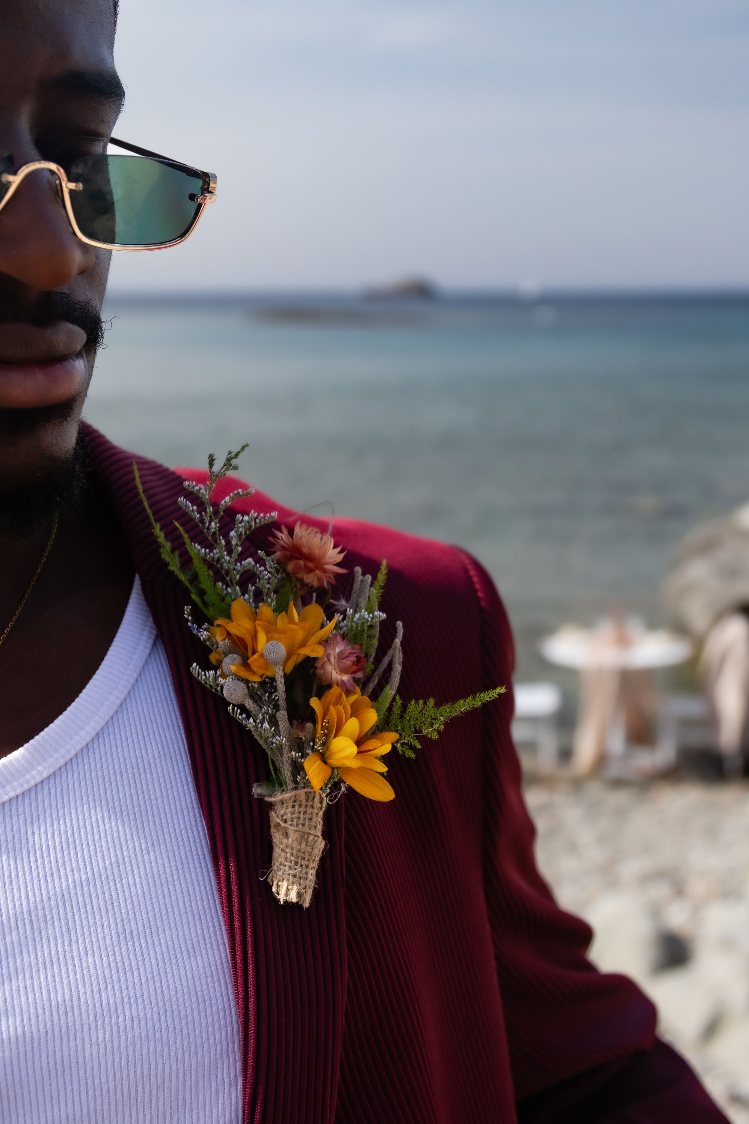 Burgundy suit jacket with floral boutonniere featuring orange lilies and wildflowers against an ocean background.