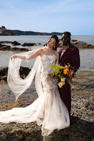 Romantic beach wedding photos with flowing white dress and burgundy details against rocky shoreline.