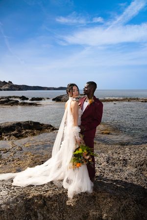 Couple shares romantic moments on a rocky beach with ocean views and a flowing white wedding dress.