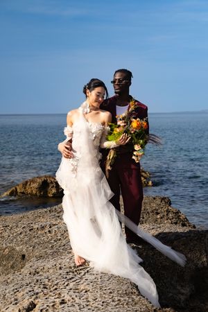 Newlywed couple embraces on rocky ocean shore with blue water and sky in background during seaside wedding photoshoot.