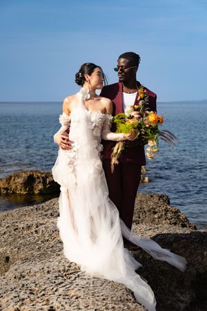 A couple in wedding attire embrace on rocky coastal shore with ocean and blue sky in background.