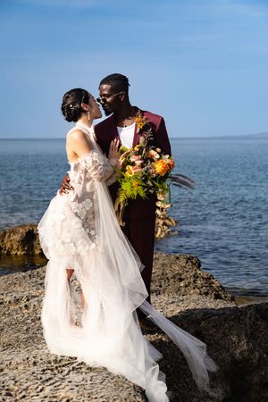 A couple in wedding attire embraces on rocky coastal cliffs overlooking a serene blue ocean on their special day.