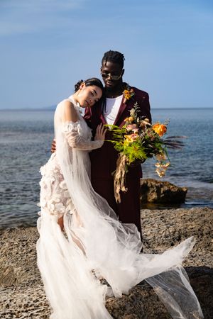 A couple in wedding attire embraces on a rocky beach with orange flowers and flowing white veil in the breeze.