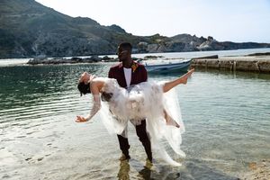 A couple in wedding attire stands in shallow turquoise waters near a stone pier with mountains in the background.