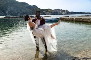A couple in wedding attire stands along a scenic rocky coastline with mountains and ocean in the background.