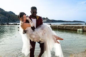 Couple in wedding attire embraces near a scenic beach with mountains and ocean views in the background.