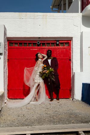 A couple in wedding attire poses playfully against a bright red garage door for artistic wedding photos.
