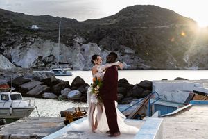 A couple shares an intimate moment at sunset on a Mediterranean harbor dock with boats and rocky cliffs in the background.