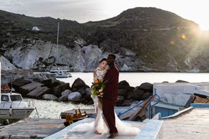 A couple embraces at sunset by a rocky harbor with boats and mountains in the background.