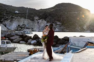 A couple shares an intimate moment at sunset on a Mediterranean dock with boats and rocky cliffs in the background.