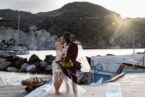 A sunset wedding portrait on a coastal dock with boats and rugged cliffs creating a dramatic backdrop.