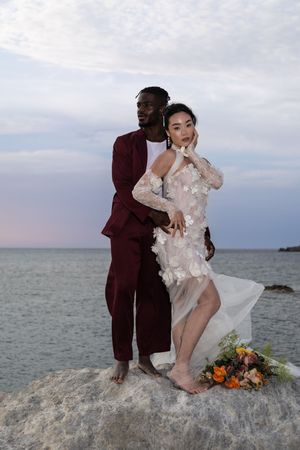 A couple shares romantic moments on a rocky beach at sunset with colorful flowers and ocean views in the background.