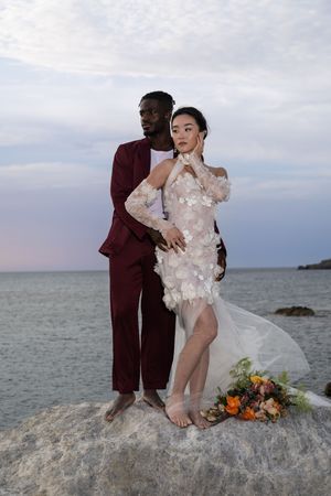 A couple poses on rocky coastal shoreline at sunset with floral bouquet and formal attire.