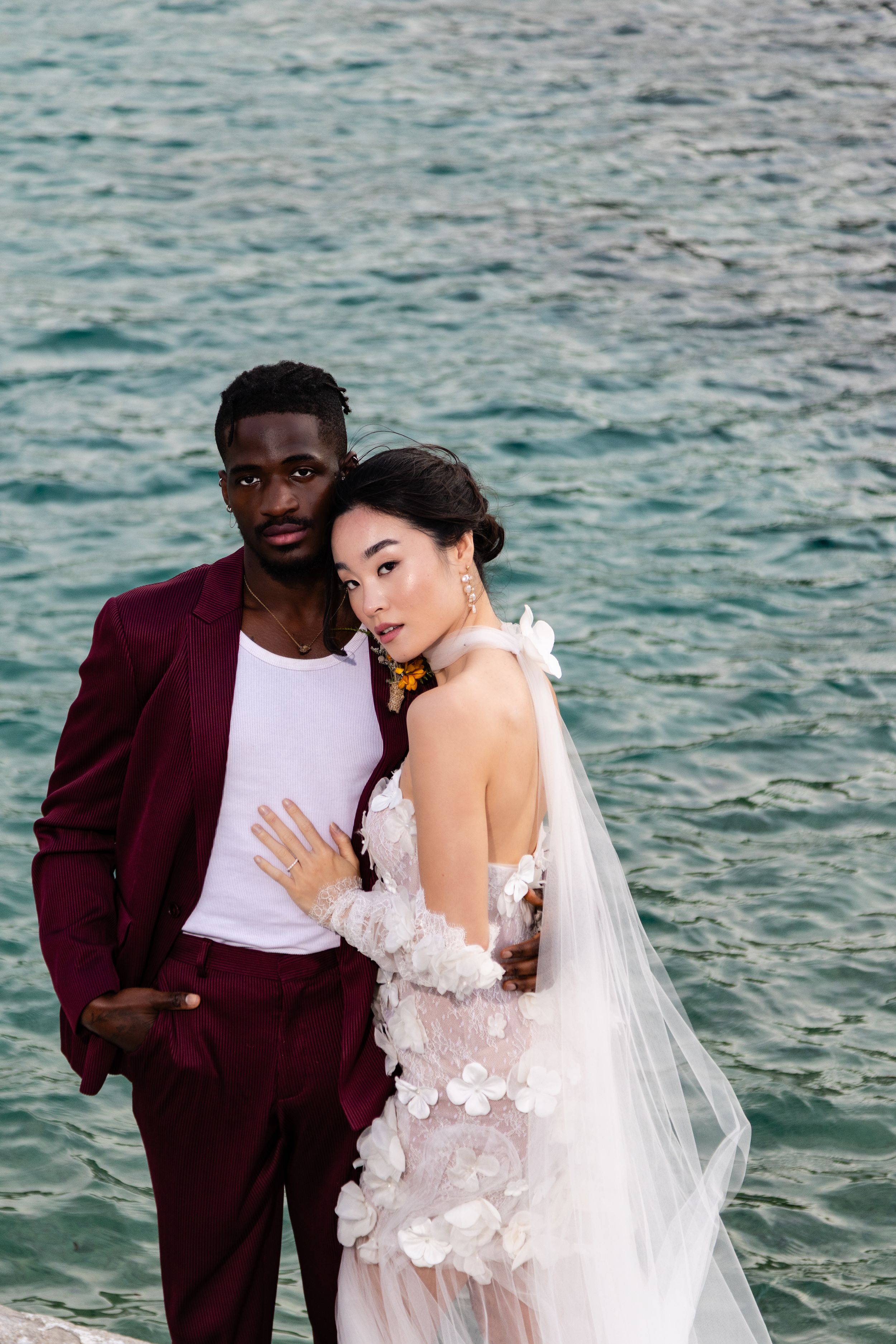 A romantic wedding photoshoot by the ocean with a couple in burgundy suit and white lace gown against turquoise waters.
