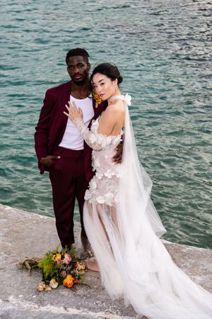 A romantic wedding couple poses together by the ocean with floral arrangements on a stone wall.