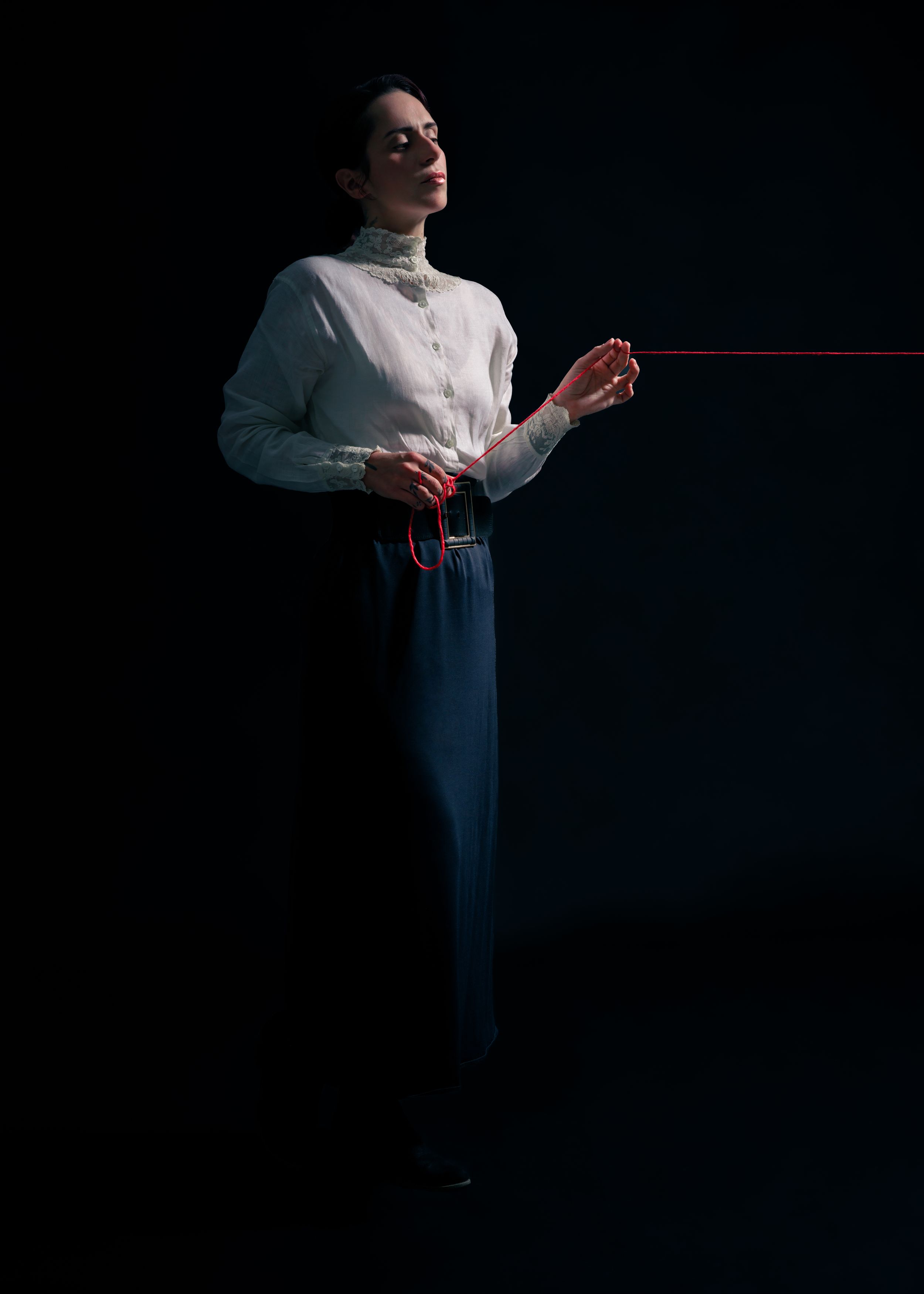 Editorial portrait of a model in a white blouse and dark trousers holding a red cord against a dark studio backdrop