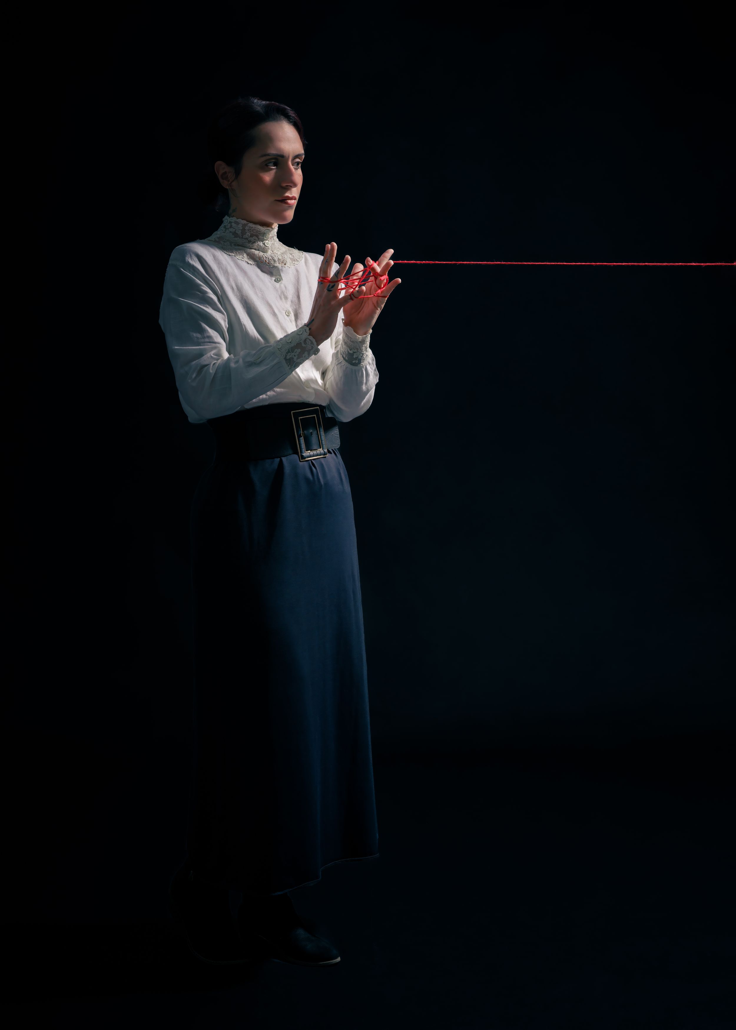 Editorial portrait of a model in a white blouse and dark trousers holding a red cord against a dark studio backdrop