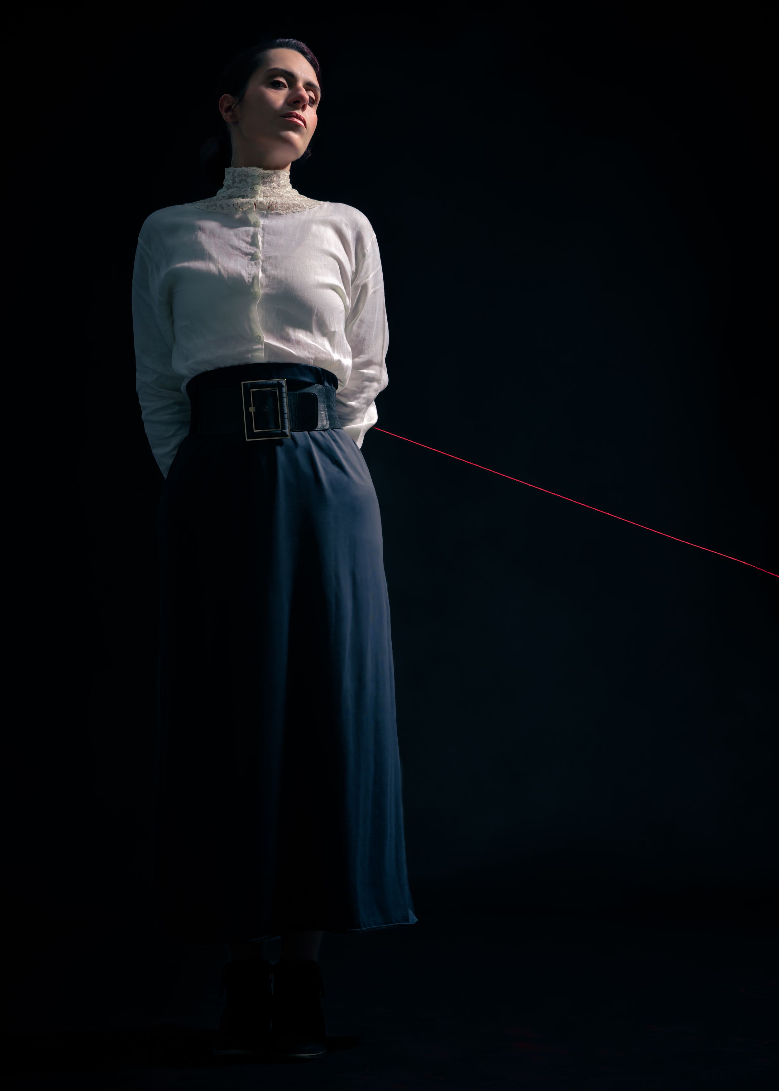 Editorial portrait of a model in a white blouse and dark trousers holding a red cord against a dark studio backdrop