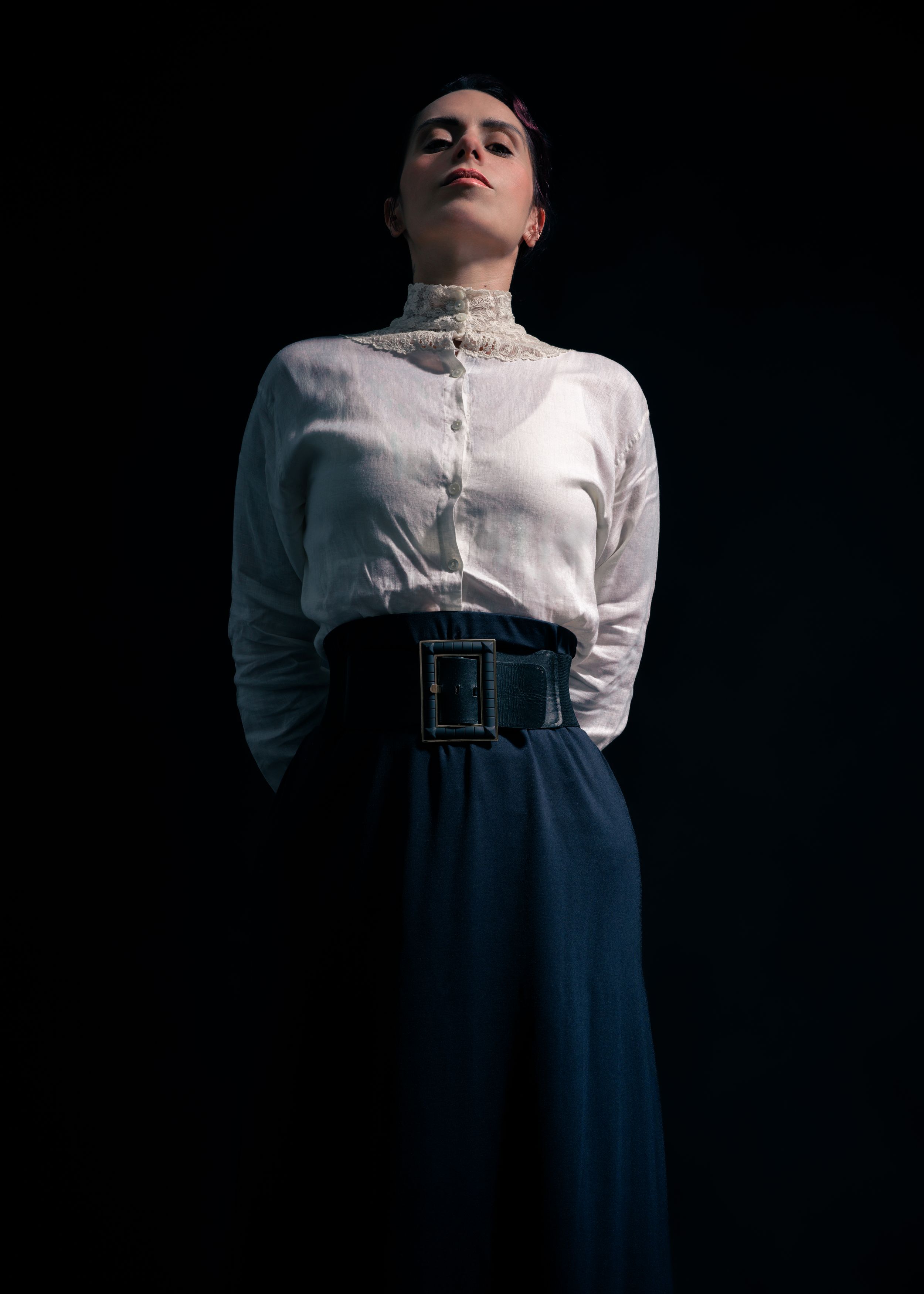 Editorial portrait of a model in a white blouse and dark wide-leg trousers against a dark studio backdrop