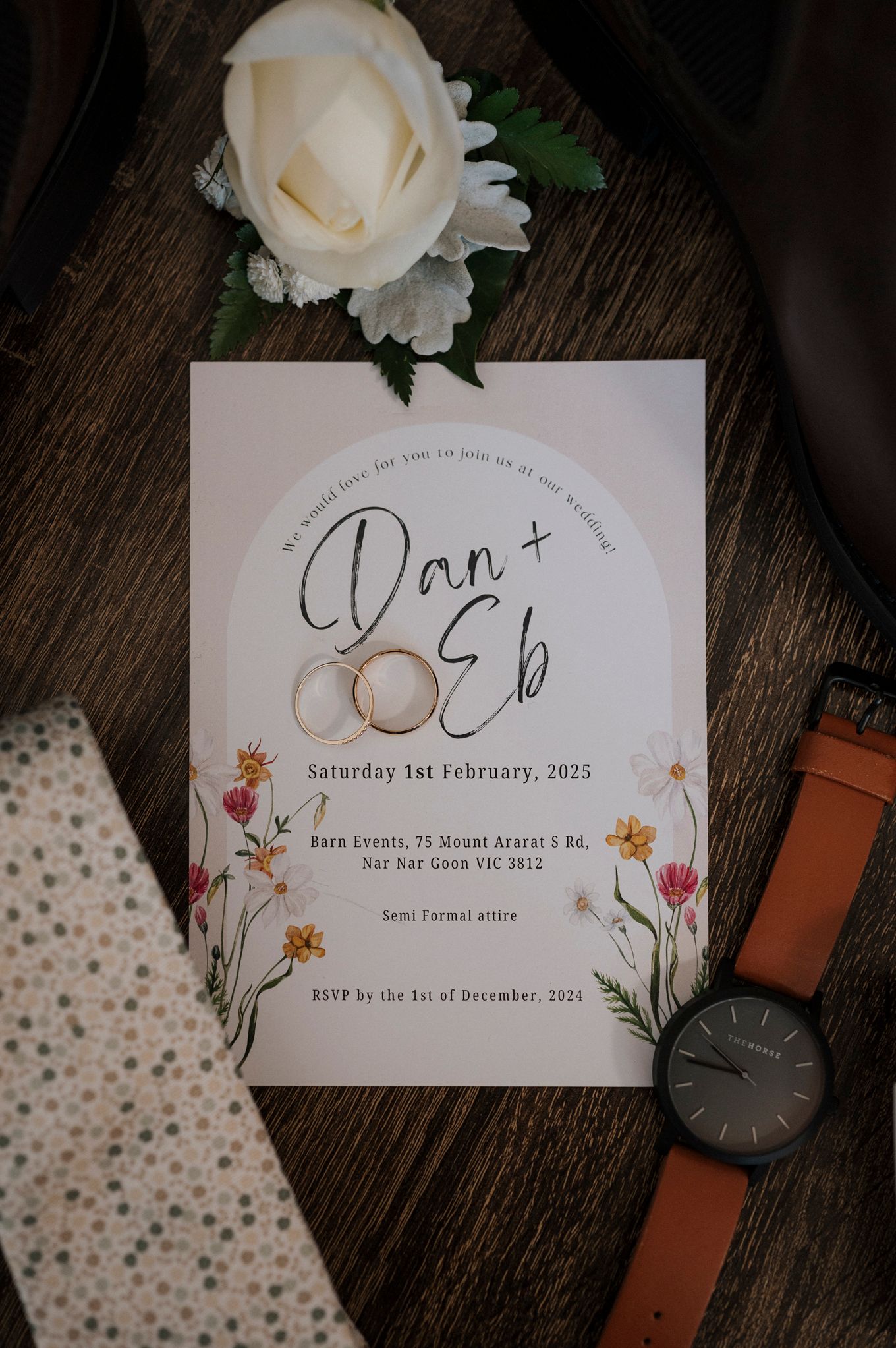 Romantic wedding invitation cards with white roses and floral details lay on a dark wooden surface next to leather accessories.