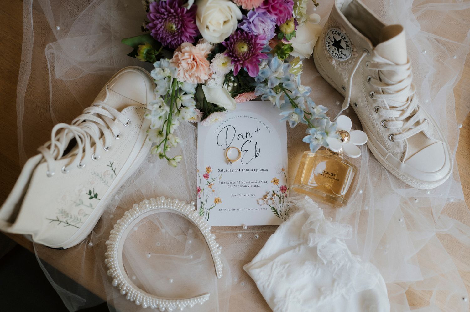 Wedding details flatlay with white sneakers, invitation card, perfume bottle, and purple floral bouquet on tulle fabric.
