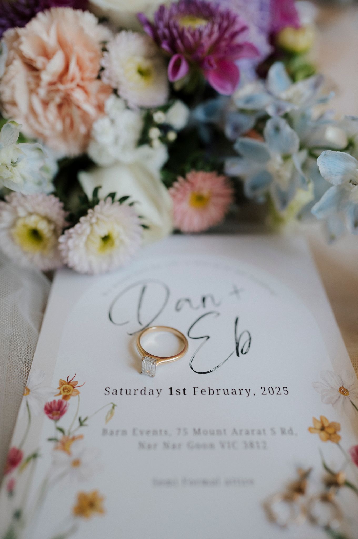 Wedding invitation for February 2023 with gold rings and pastel flower arrangement featuring carnations and daisies.