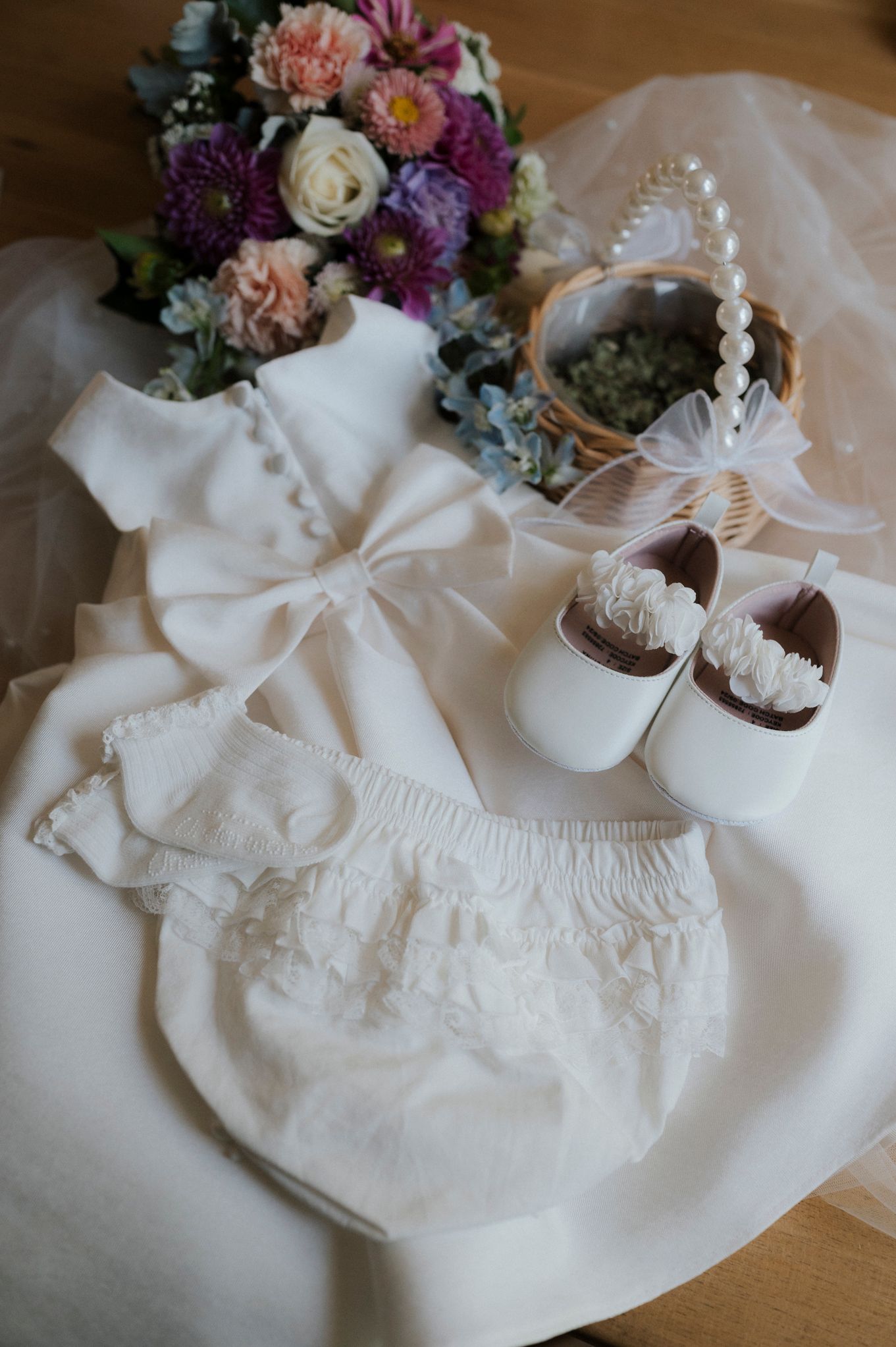 Delicate white baby dress with matching accessories and floral arrangements displayed on wooden surface.