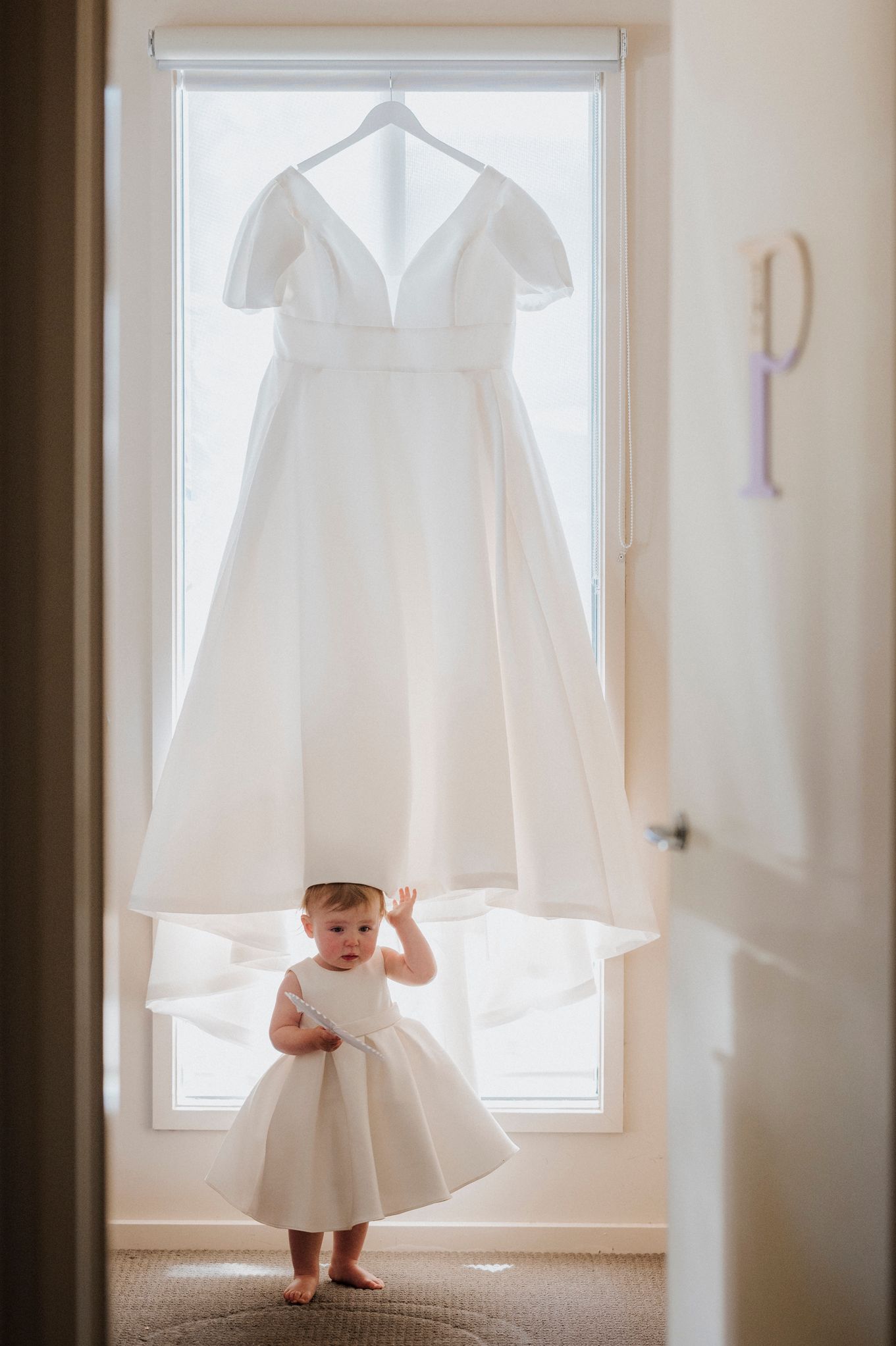 A small child in a white dress stands beneath a hanging white wedding gown in front of a bright window.