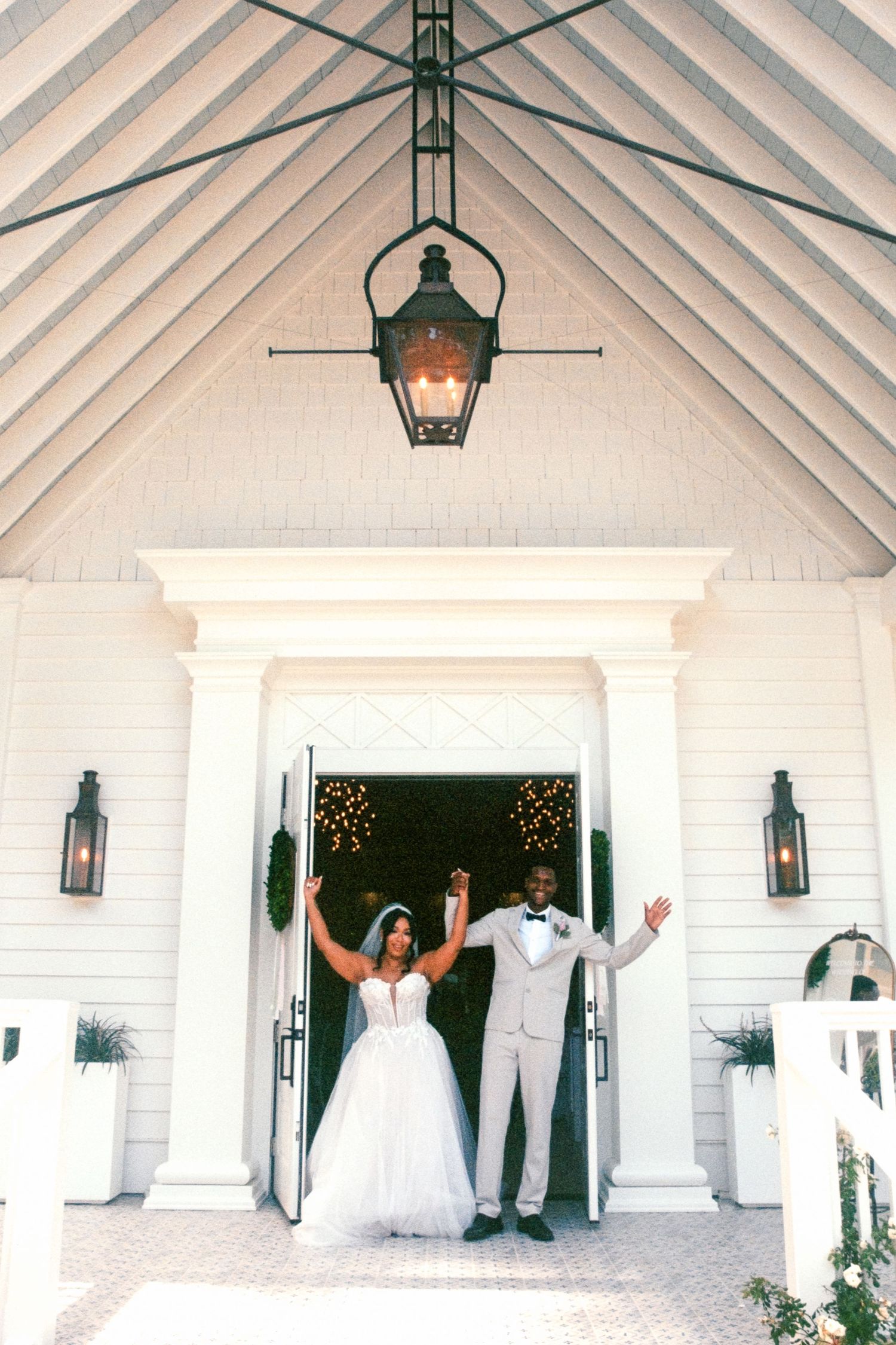 A couple celebrates at an elegant white wedding chapel entrance with antique lanterns and twinkle lights in the background.