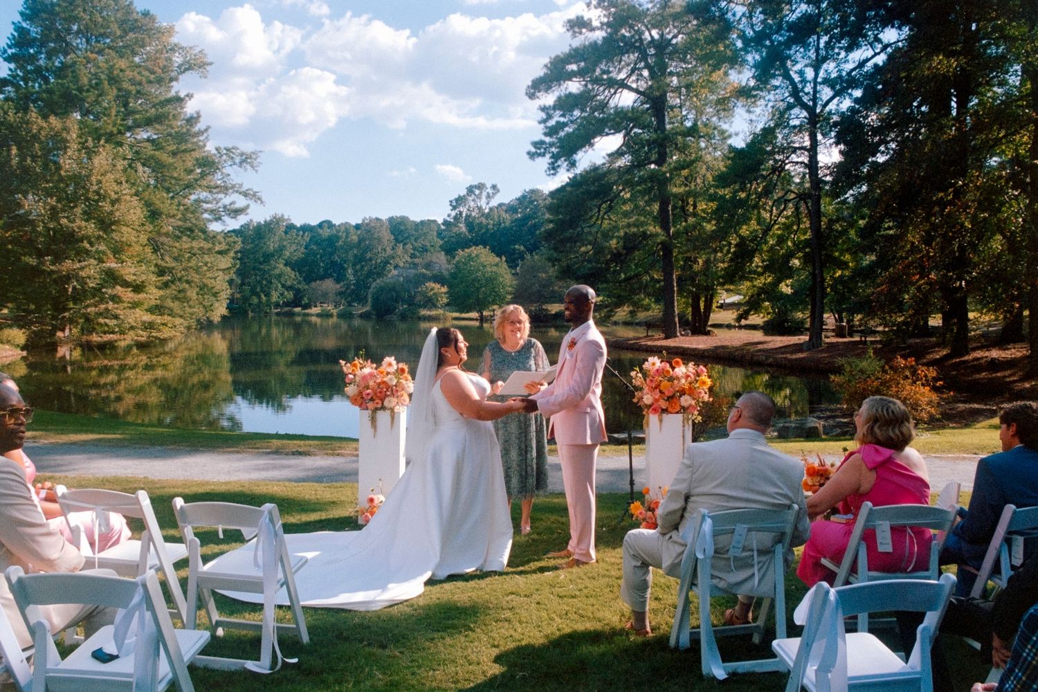 Wedding party dressed in white celebrates with raised arms on a lakeshore dock during sunset.
