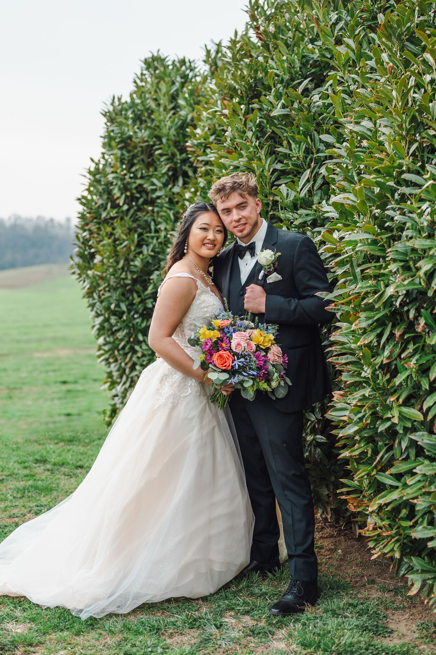A couple poses outdoors next to tall hedges on their wedding day, with the bride holding a vibrant multicolored bouquet.